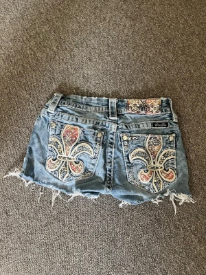 Blå jeansshorts med broderade fickor - Säljer ett par blå jeansshorts från Miss Me med slitna kanter och stora broderade detaljer på bakfickorna. Shortsen har dekorativa nitar och ett färgglatt mönster på fickorna samt bältet. Perfekta för sommaren och ger en cool, avslappnad look.