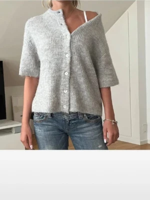 Grå ribbstickad kofta med knappar - Mysig grå ribbstickad kofta med korta ärmar och vita knappar framtill. Koftan har en avslappnad passform och är perfekt att slänga över en topp. Enkel och stilren design som passar till jeans eller kjol.