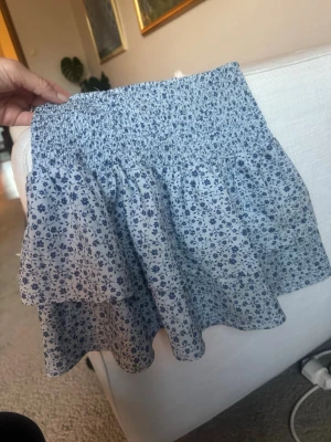 Blommig kortkjol från Gina Tricot - Supersöt kortkjol från Gina Tricot med blått blommönster på ljus botten. Kjolen har smock i midjan och volangdetaljer som ger en luftig och somrig känsla.