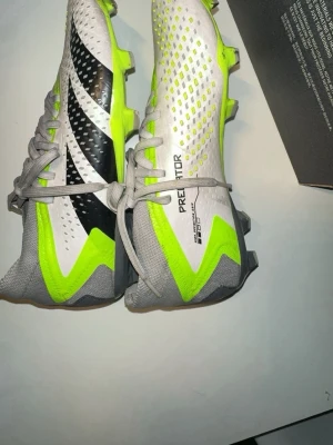 Adidas elite Predator fotbollsskor vit/neon - Säljer ett par Adidas Predator fotbollsskor i vitt, grått, svart och neon-gult. Skorna har snörning, mönstrad ovandel och tydliga Adidas-loggor. Designen är modern med skarpa färgkontraster och greppvänliga dobbar för spel på gräs.
