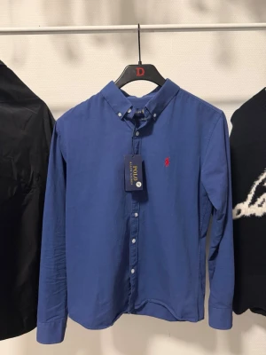 Blå skjorta från Polo Ralph Lauren - Klassisk blå skjorta från Polo Ralph Lauren med button-down krage och röd broderad logga på bröstet. Skjortan har långa ärmar och vita knappar framtill. Perfekt för en stilren och tidlös look.