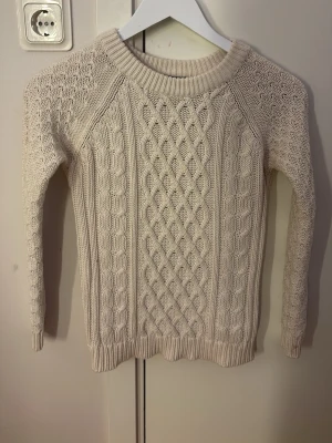 Beige kabelstickad tröja från Vila - Snygg beige kabelstickad tröja från Vila med rund halsringning och ribbade muddar. Tröjan har ett klassiskt flätmönster framtill och är långärmad. Perfekt för lager-på-lager och passar till många olika stilar.