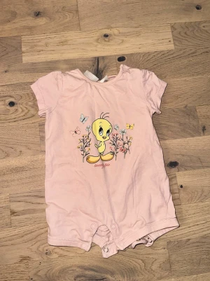Rosa body med Tweety-motiv - Söt rosa body med korta ärmar och tryck av Tweety Pie bland blommor och fjärilar. Praktiska knappar i grenen för enkel på- och avklädning. Perfekt för dig som gillar gulliga och lekfulla motiv.
