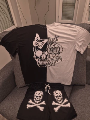 Svartvitt set med dödskallemotiv M/S - Säljer ett coolt svartvitt set med t-shirt och shorts. T-shirten har ett stort dödskalletryck med rosor och fjäril på framsidan, och är delad i svart och vitt. Shortsen är svarta med dödskalle och korsade ben på framsidan. Perfekt för dig som gillar edgy stil!