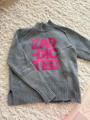 zadig voltaire tröja - Cool grå stickad tröja med hög krage och stora rosa bokstäver 'ZAD-DICTED' på bröstet. Tröjan har ribbstickade muddar och nederkant samt slitsar på sidorna. Perfekt för dig som gillar statement-plagg och vill sticka ut.