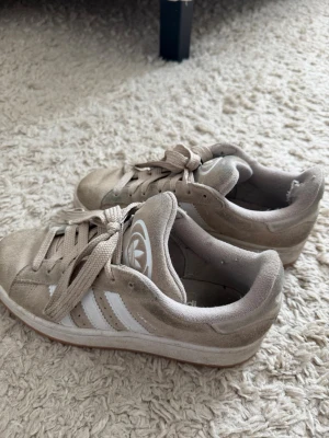 Beige Adidas sneakers med mocka - Säljer ett par beige Adidas sneakers i mocka med klassiska vita ränder på sidorna och Adidas-logga på plösen. Skorna har låga skaft och beige snörning. Yttersulan är i gummi och designen är stilren och enkel. Lite slitna, går och tvätta av smutset.