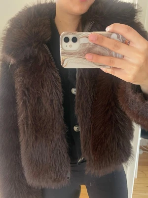 Mörkbrun fluffig pälsjacka - Säljer en mörkbrun pälsjacka med bred krage och dragkedja framtill. Jackan har en kortare, boxig passform och är gjord i ett mjukt, fluffigt material som ger en lyxig känsla. Perfekt för att hålla värmen under kalla dagar.