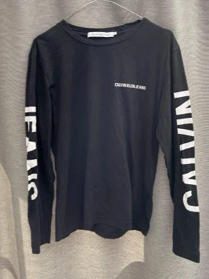 Svart långärmad topp från Calvin Klein - Svart långärmad topp från Calvin Klein Jeans med vit logotyp på bröstet och stora vita tryck på ärmarna och ryggen. T-shirten har en normal passform och är gjord i mjuk bomull, perfekt för en cool och avslappnad look.
