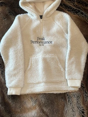 Vit teddyhoodie från Peak Performance - Mysig vit hoodie i teddy-material från Peak Performance med broderad logga på bröstet. Halvlång dragkedja framtill, stor huva och känguruficka. Perfekt för kalla dagar och chill outfits.