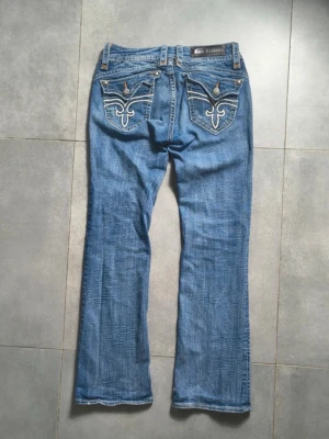 Blå bootcut jeans från Rock Revival - Snygga blå bootcut jeans från Rock Revival med dekorativa broderier och detaljerade bakfickor. Jeansen har en klassisk femficksmodell, slitningar framtill och kontrastsömmar. Perfekta för dig som gillar en cool och avslappnad stil.