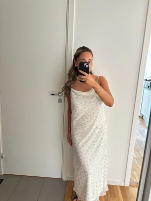 Vit långklänning med blommigt mönster - Söt vit långklänning med tunna axelband och små blå blommor. Klänningen har en avslappnad passform och mjukt fallande tyg, perfekt för varma dagar. Lätt och luftig känsla med en enkel look.
