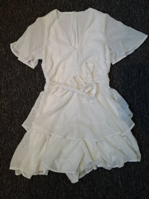 Vit prickig jumpsuit med volanger - Supersöt vit klännung med korta ärmar, omlott-look och knytband i midjan. Tyget har ett diskret prickigt mönster och volangdetaljer längs benen och ärmarna. Perfekt för sommarens alla äventyr! Det är också in byggda shorts