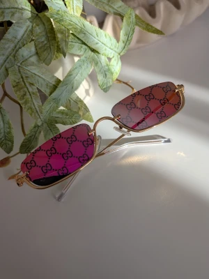 GUCCl SOLGLASÖGON  - Unika solglasögon i en djärv design med rödrosa tonade glas och guldfärgade detaljer. Glas: Rektangulära linser med ett heltäckande svart monogram-mönster som skapar en iögonfallande effekt. Bågar: Slank konstruktion i guldfärgad metall med en klassisk näsbrygga och diskreta skalmar. Stil: En modern retro-look med stark karaktär, inspirerad av tidigt 2000-tal och streetwear-mode. Detaljer: Transparenta ändtippar på skalmarna för ökad komfort.