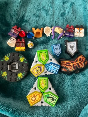 Lego Nexo Knights tillbehör & sköldar (Offer) - Samling med Lego Nexo Knights tillbehör, inklusive flera sköldar med olika motiv, hjälmar, huvuddelar och rustningar. Färgerna är blandade med inslag av grönt, blått, svart, orange och gult. Perfekt för att bygga egna figurer eller komplettera din samling.