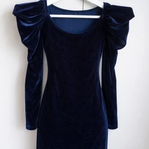 Marinblå sammetsklänning Gina Tricot - Marinblå sammetsklänning från Gina Tricot med långa ärmar och markerade puffaxlar. Klänningen har en figurnära passform och rund halsringning, perfekt för dig som vill sticka ut med en elegant och trendig look.