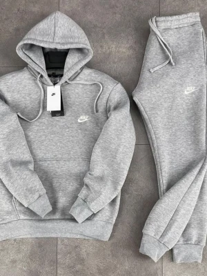 Grått Nike loungewear-set - Mysigt grått loungewear-set från Nike med hoodie och matchande byxor. Hoodien har dragsko och magficka, byxorna har resår och dragsko i midjan. Båda plaggen har Nike-logga broderad i vitt. Perfekt för chill dagar hemma.
