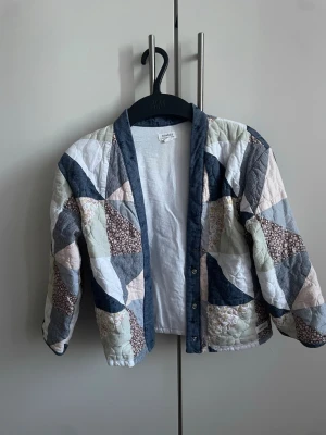 Patchwork cardiganjacka från Newbie - Patchwork cardiganjacka från Newbie i storlek 122. Jackan har ett unikt lapptäcksmönster i blått, vitt, beige och brunt med olika tyger. Den är quiltad och har tryckknappar framtill samt mjuk bomullskänsla. Perfekt för dig som gillar retrostil och mysiga lager.