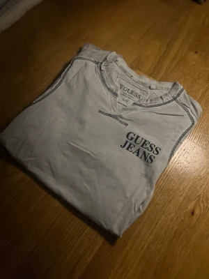 Vit t-shirt från Guess Jeans -  T-shirt från Guess Jeans med blå logotyp på bröstet och dekorativa sömmar. Klassisk passform och rund halsringning. Perfekt för dig som gillar stilrena plagg med diskret branding.
