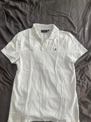 Vit pikétröja från Morris - Säljer en klassisk vit pikétröja från Morris med krage och korta ärmar. Tröjan har en liten broderad logotyp på bröstet och knappar vid halsen. Perfekt för en clean och stilren look.