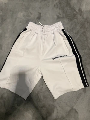 Palm Angels vita shorts med svarta ränder - Palm Angels vita shorts med svarta ränder längs sidorna och logotyp på ena benet. De har elastisk hög midja och sidofickor. Materialet är polyester och passformen är loose, perfekt för en avslappnad och cool stil.