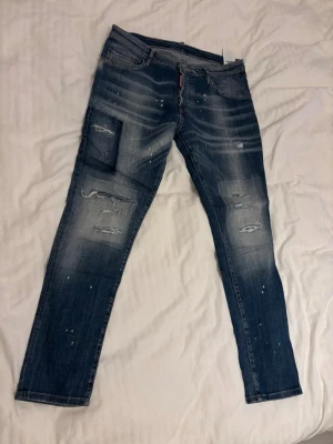 Blå slitna jeans från Dsquared2 - Säljer ett par blå jeans från Dsquared2 med slitningar. Jeansen har en smal passform, klassiska fem fickor och vita färgstänk för en trendig look. lite slitet på patchet där bak annars mycket fina jeans. För funderingar mätningar osv är det bara o skriva.