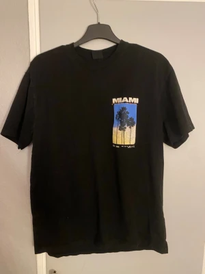 Svart Miami t-shirt med palmtryck - Cool svart t-shirt med Miami-tryck och palmer på både fram- och baksidan. Motivet har färgskiftningar i blått, rosa och beige som ger en snygg kontrast. Perfekt för dig som gillar streetstyle och grafiska prints.