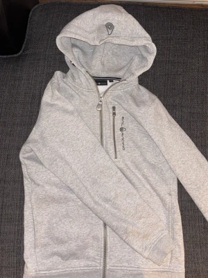 Ljusgrå zip hoodie från Sail Racing - Ljusgrå hoodie med dragkedja från Sail Racing. Hoodien har en broderad logga på huvan och tryckt logga på bröstet. Praktisk bröstficka med dragkedja och två sidofickor. Mjuk bomullsblandning och ribbade muddar vid ärmar och nederkant.