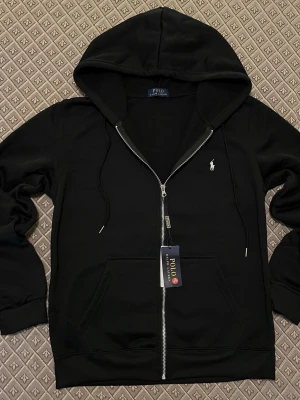 Svart hoodie från Polo Ralph Lauren - Snygg svart hoodie från Polo Ralph Lauren med dragkedja framtill och klassisk huva med dragsko. Ikoniska vita loggan broderad på bröstet. Tillverkad i mjukt bomullsmaterial och har två fickor framtill. Perfekt för en avslappnad och stilren look.