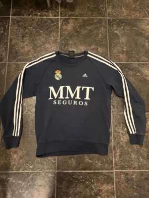 Blå Real Madrid Adidas sweatshirt 2000-talet - Mörkblå sweatshirt från Adidas med Real Madrid-logga och MMT Seguros-tryck på bröstet från omkring 2000-talet. Tröjan har vita ränder längs ärmarna och rund hals.