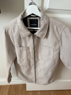 Overshirt från Jack & Jones - Stilren beige jacka från Jack & Jones med två bröstfickor och dragkedja framtill. Klassisk krage och raka ärmar. Tillverkad i mjuk bomull som passar perfekt till vår och höst.
