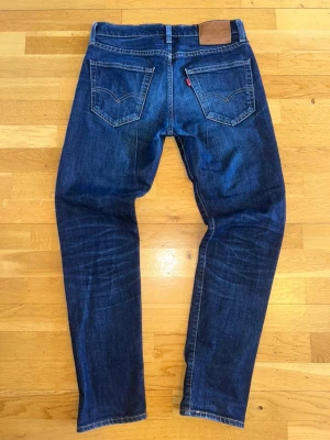 Levi's 512 - Säljer ett par klassiska mörkblå Levi's jeans med raka ben och fem fickor. Jeansen har snygga slitningar och den ikoniska läderpatchen bak i midjan. Perfekta för dig som gillar tidlös denimstil.
