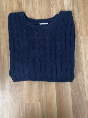 Mörkblå kabelstickad tröja - Mysig mörkblå kabelstickad tröja med rund halsringning och ribbade muddar vid ärmslut och nederkant. Klassisk design som passar perfekt till jeans eller kjol. Materialet känns mjukt och bekvämt mot huden.