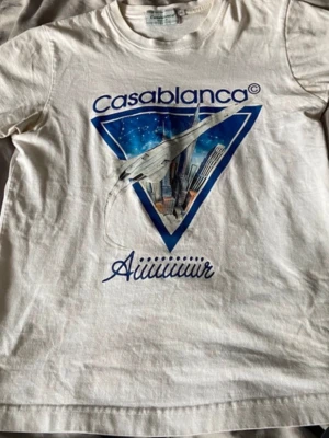 Vit Casablanca t-shirt med tryck - Vit t-shirt från Casablanca med stort grafiskt tryck framtill i blått och vitt, föreställande en Concorde och stadssilhuett. Rund halsringning och klassisk passform. Perfekt för dig som gillar unika statement-plagg.