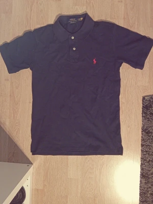 Marinblå pikétröja från Ralph Lauren - Klassisk marinblå pikétröja från Ralph Lauren med korta ärmar och två knappar vid kragen. Ikoniska röda loggan broderad på bröstet. Perfekt basic till garderoben och lätt att matcha med allt. Säljes då den är för liten. OBS!! Priset är inte hugget i sten så tveka inte att höra av dig!!
