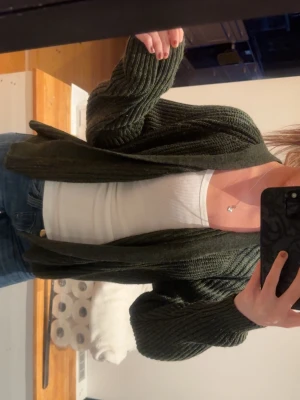 Mörkgrön stickad kofta - Mysig mörkgrön stickad kofta med öppen front och ribbade detaljer. Koftan har en avslappnad passform och långa ärmar, perfekt att slänga över en topp eller t-shirt. Passar till jeans eller kjol för en chill look.