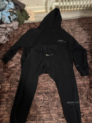 Svart Essentials hoodie med huva - Säljer en svart hoodie från Essentials med huva och diskret logga på bröstet och ärmen. Hoodien har en klassisk passform och är gjord i mjukt material som känns skönt mot huden. Perfekt för dig som gillar streetwear och enkel stil.