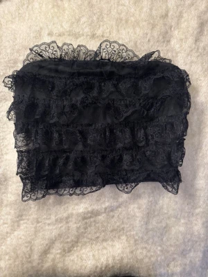 Svart spets bandeau tubtopp - Snygg svart bandeau tubtopp med flera lager volangig spets och transparenta spetsdetaljer. Toppen är axelbandslös och har en feminin, croppad passform som passar perfekt till höga jeans eller kjol.