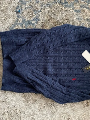 Mörkblå kabelstickad tröja Polo Ralph Lauren - Mörkblå kabelstickad tröja från Polo Ralph Lauren med klassisk röd broderad logga på bröstet. Tröjan har långa ärmar, ribbade muddar och rund hals. Perfekt för dig som gillar stilrena och tidlösa plagg i mjukt material.