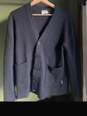 Mörkblå kofta från Lindbergh - Mysig mörkblå kofta från Lindbergh i lammull med comfort fit. Koftan har djup V-ringning, knäppning med fyra knappar och två stora fickor framtill. Ribbstickade muddar vid ärmslut och nederkant ger en klassisk look.