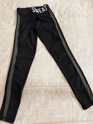 Svarta tights med färgade ränder - Snygga svarta träningstights med elastisk midja där det står 'SWEAT NOW'. Ena sidan har gröna ränder och andra sidan har rosa ränder längs benen. Perfekta för gymmet eller löpning. Materialet är stretchigt och andas bra.