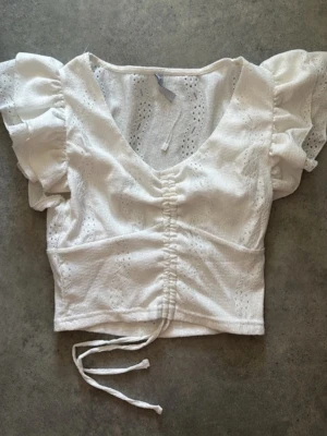 Vit broderad croppad blus med snörning - Superfin vit blus med broderade detaljer och puffiga korta ärmar. Toppen är croppad och har snörning framtill som ger en snygg rynkad effekt. Perfekt till sommaren och har en v-ringad halsringning för en trendig look.