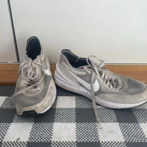 Nike gråvita sneakers med mesh - Säljer ett par gråvita Nike sneakers med klassisk vit swoosh på sidan. Skorna har meshpaneler för extra andningsförmåga och detaljer i mocka och syntet. Lågt skaft och snörning framtill. Perfekta för en sportig och avslappnad stil.