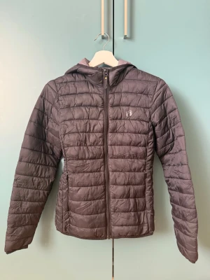 Jacka  - Snygg pufferjacka med huva och dragkedja framtill. Jackan har quiltad design, två sidofickor med dragkedja och diskret logga på bröstet. Perfekt för kalla dagar och riktigt skön att bära.