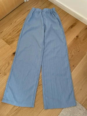 Ljusblå vida linnebyxor - Säljer ett par ljusblå linnebyxor med avslappnad passform och resår i midjan. Byxorna har raka, vida ben och är tillverkade i ett tunt, luftigt material som passar perfekt för varma dagar. Inga synliga fickor eller detaljer. 51 cm i längden 