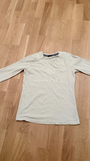 Beige långärmad topp från Gant - Snygg beige långärmad topp från Gant med rund halsringning och stort vitt logotryck framtill. Tillverkad i mjuk bomull som känns skön mot huden. Perfekt att styla med jeans eller kjol för en clean look. Innan jag fraktar så ser jag till att jag har strykt den!