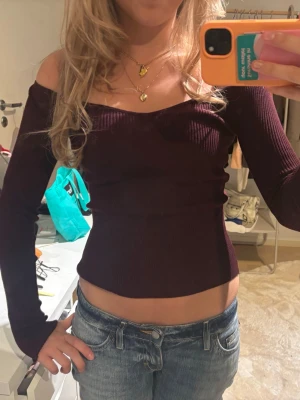 Mörklila offshoulder ribbad topp - Trendig mörklila offshoulder topp med lång ärm och ribbad struktur. Sitter tight och har en snygg croppad passform som framhäver midjan. Perfekt för dig som gillar en stilren men ändå edgy look.