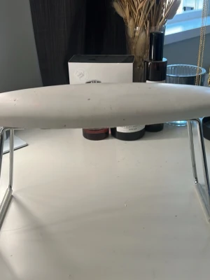 Vit nageldyna med metallben - Nageldyna i avlång, oval form med vit ovansida och grå undersida i mjukt tyg. Dynan har stabila metallben som ger bra stöd när du gör naglar. Perfekt för att vila händerna på under nagelbehandlingar hemma eller i salong. Nypris 299