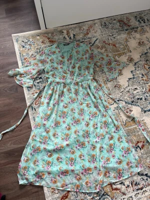 Blommig långklänning från mbyM - Ljusblå långklänning från mbyM med blommigt mönster i rosa, orange och lila. Klänningen har v-ringning, knappar framtill, avtagbart bälte i midjan och transparenta, vida ärmar. Perfekt för vår och sommar!