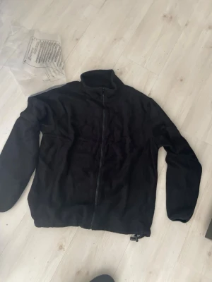 Svart fleecejacka med dragkedja - Mjuk och enkel svart fleecejacka med hel dragkedja framtill och hög krage. Perfekt för lager-på-lager och chill dagar. Jackan har långa ärmar och justerbar nederkant med dragsko. Passar dig som gillar basic och funktionellt.