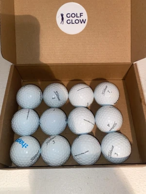 Titlesit PRO V1 A/B-Klass - Säljer ett set med tolv vita golfbollar från Titleist, levereras i en enkel brun kartong med Golf Glow-klistermärke på insidan av locket. Klassisk rund form och tydlig märkning på varje boll.